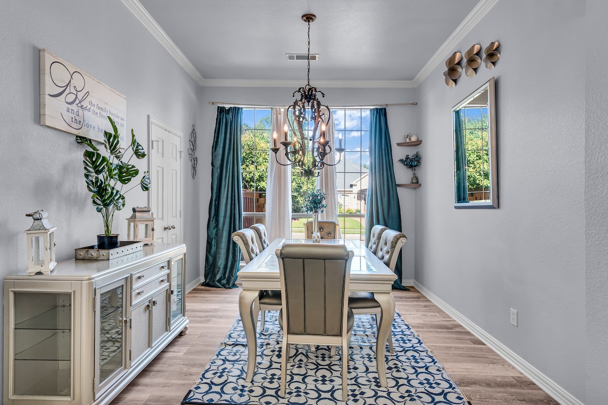 A bright dining area is presented with a large white table surrounded by elegant chairs. Natural light filters through large windows adorned with blue curtains, creating an inviting atmosphere. A decorative chandelier hangs above, while a plant adds a touch of greenery.