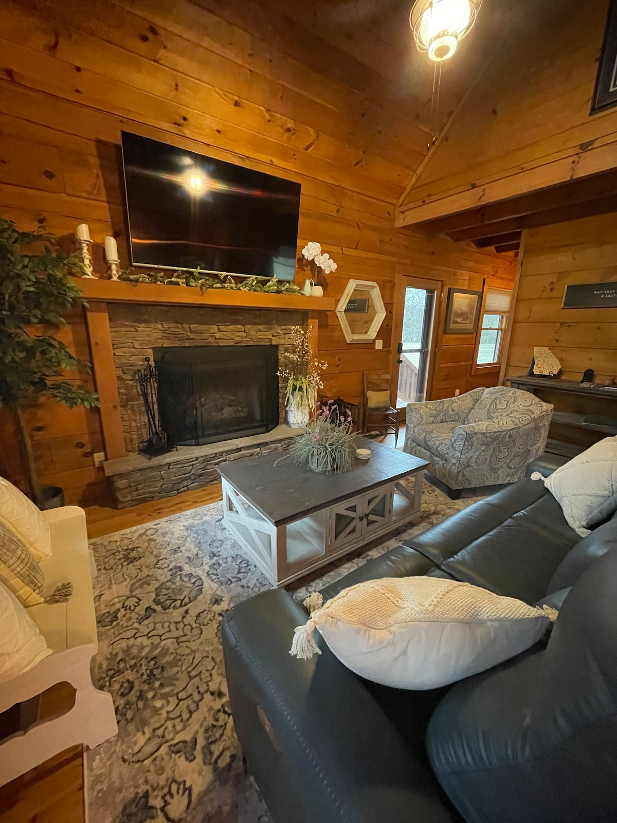 The cozy living room features a stone fireplace beneath a mounted television. A plush dark seating arrangement is positioned for comfort, accompanied by a decorative coffee table and a patterned area rug. Natural light enters through nearby windows, enhancing the warm wooden interior.