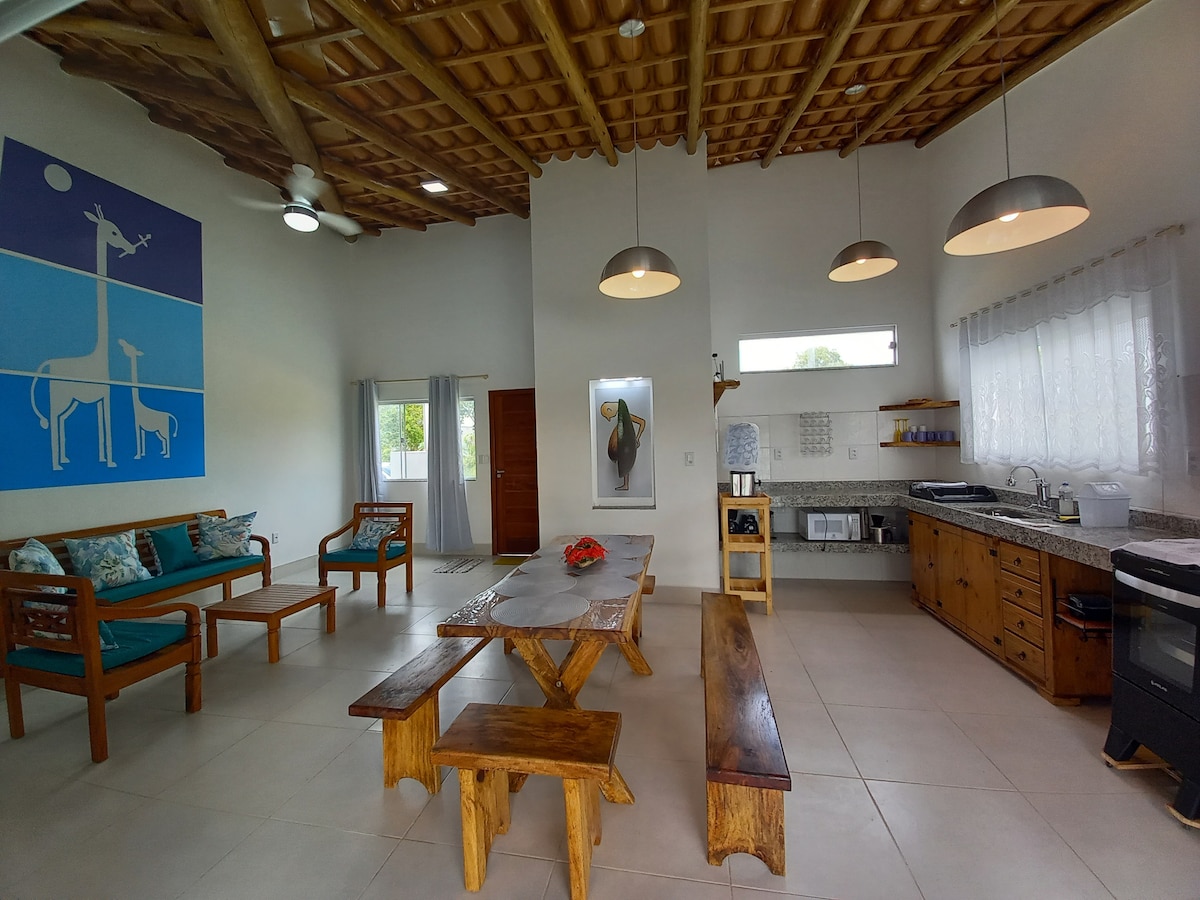 A spacious living area features high ceilings adorned with wooden beams, accommodating a dining table made of natural wood and a cozy seating arrangement. A large window allows natural light, and a blue wall art piece depicting giraffes adds a playful touch.