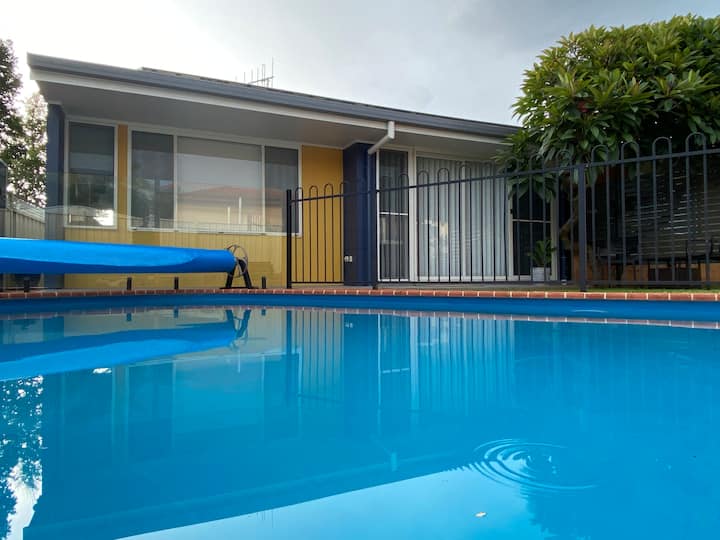 A backyard pool with a blue pool cover roller sits in front of a single-story house with large windows and a sliding door. A black metal fence surrounds the pool area, and a lush tree adds greenery to the space