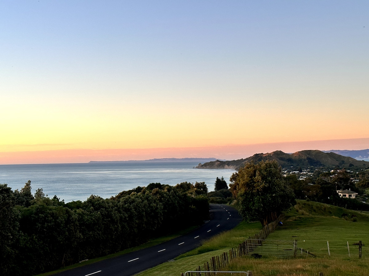 An expansive view of the ocean stretches beneath a soft gradient sky, transitioning from warm colors to twilight tones. Lush greenery frames the foreground, while a winding road gently curves towards the horizon, leading to distant hills and coastal scenery.