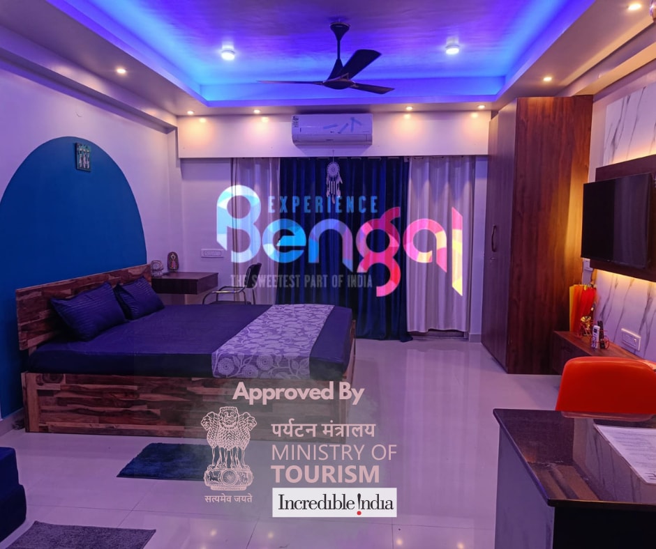 A spacious studio is illuminated by soft blue lighting. A large bed with dark bedding is visible, accompanied by a wooden accent wall. A seating area with colorful chairs and a sleek TV unit enhances the modern design. Bright curtains frame the window, adding to the inviting ambiance.