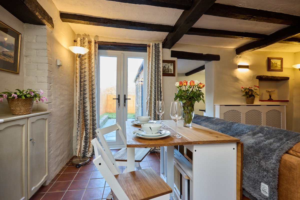 A dining area is presented with a wooden table set for two, featuring dishes and glasses. Light streams through double doors that open to a garden. Textured curtains frame the doors, while decorative wall art and potted plants add subtle charm to the space.