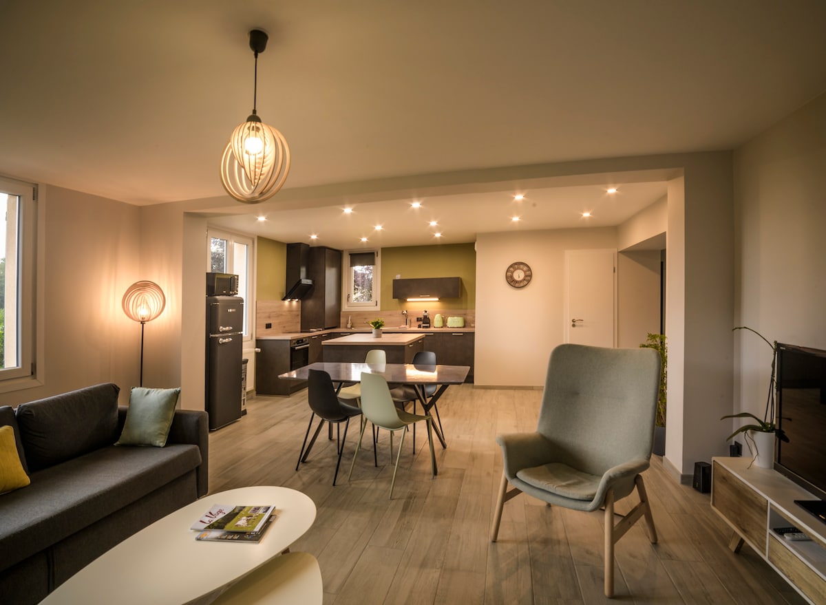 An open living space is presented, featuring a cozy sofa and a modern armchair. A dining table with colorful chairs sits adjacent to the well-equipped kitchen, illuminated by soft ceiling lights. Large windows allow natural light to brighten the area.
