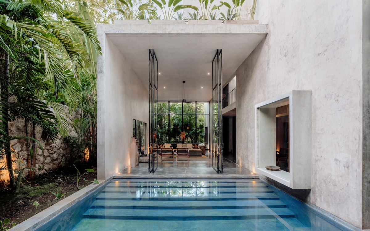 An inviting outdoor area showcases a private pool with a sleek blue tiled surface, reflecting the surrounding lush greenery. The adjacent indoor space features floor-to-ceiling windows, allowing natural light to illuminate the living area, which is designed for relaxation and connection with nature.