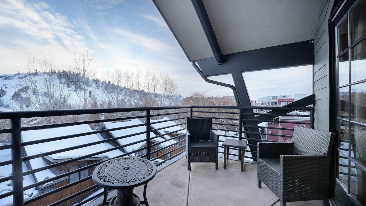 A private balcony is presented with seating arrangements, including two chairs and a small table. Snow-covered rooftops are visible below, with a view of the hills in the distance. The soft morning light highlights the serene outdoor space.