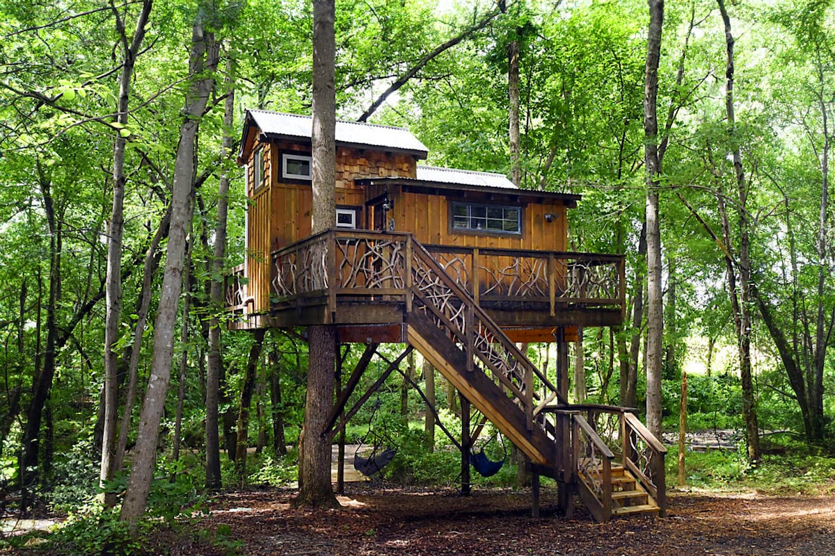 An elevated treehouse is surrounded by lush green trees, showcasing a rustic wooden exterior. A staircase leads to a spacious deck with decorative railing, providing a comfortable area to relax. Below, a swing hangs from a nearby tree, adding to the tranquil outdoor setting.