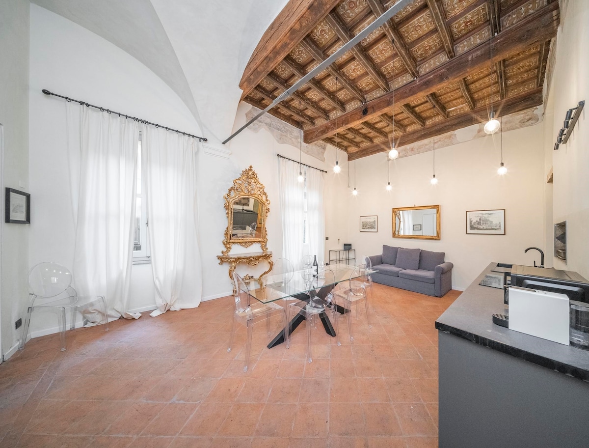 An elegantly furnished living area showcases a spacious sofa and a glass dining table surrounded by transparent chairs. The room features high ceilings with wood beams and natural light streaming through large curtains. A decorative mirror and contemporary lighting elements enhance the ambiance.