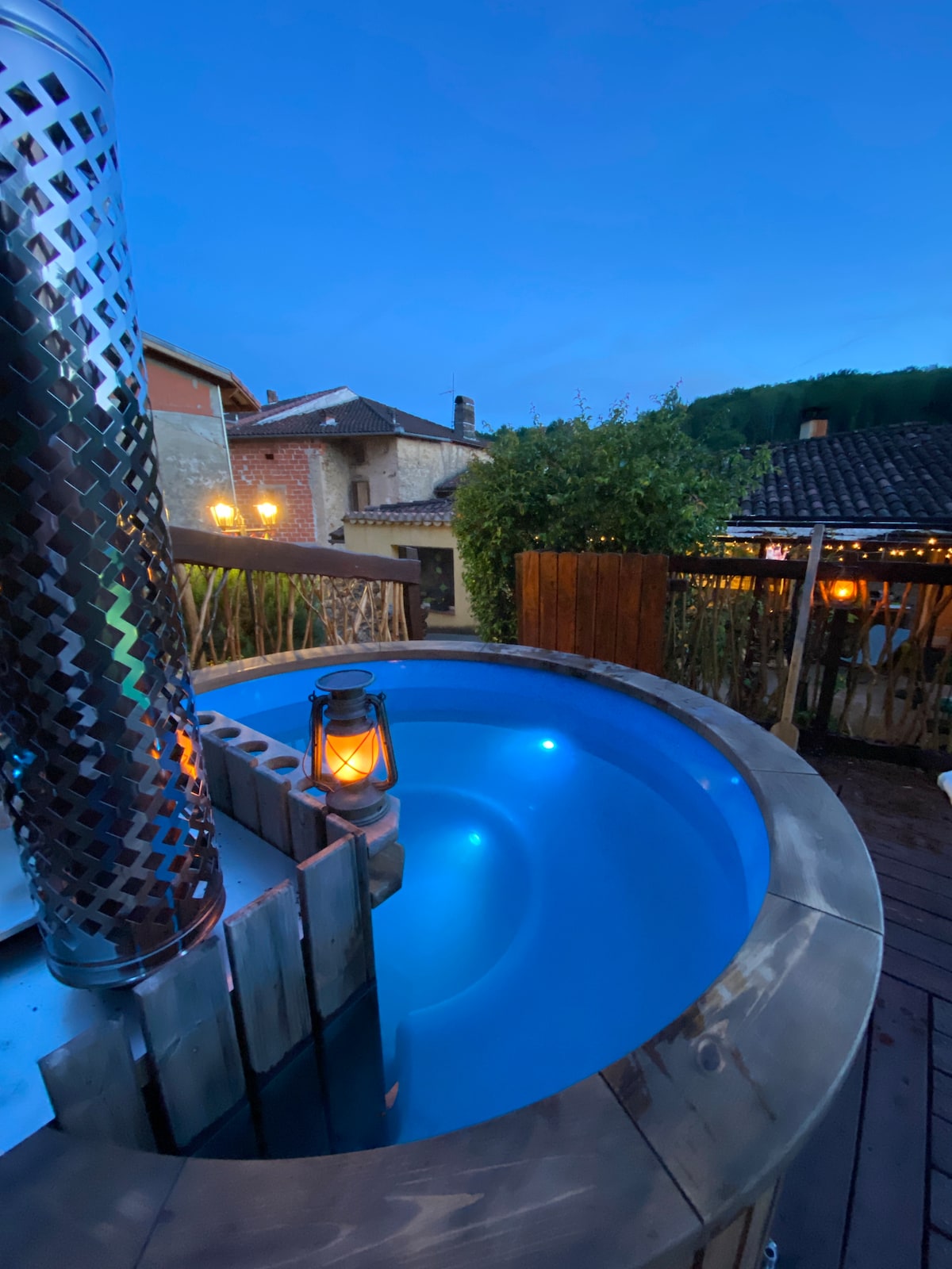 A circular wooden hot tub is featured on a wooden deck, surrounded by lush greenery and charming buildings. The soft blue water is illuminated, creating a calming atmosphere. A lantern provides gentle light, enhancing the tranquil evening setting.