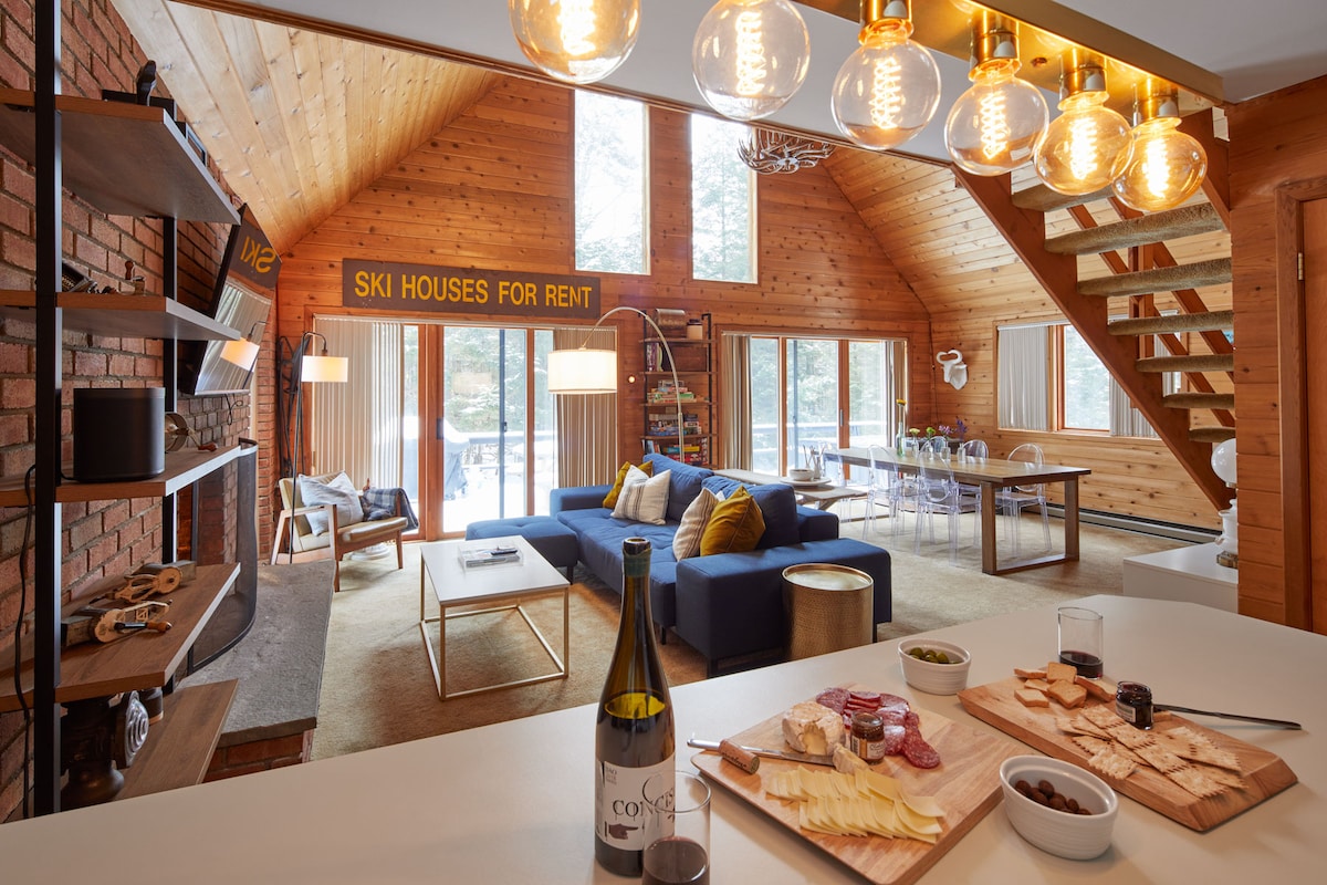 An open-concept living area features a deep blue sectional sofa adorned with yellow accent pillows. A wooden staircase leads to the upper level, while large windows provide views of the surrounding nature. A dining table is set for gatherings, with a charcuterie board displayed on a kitchen island.