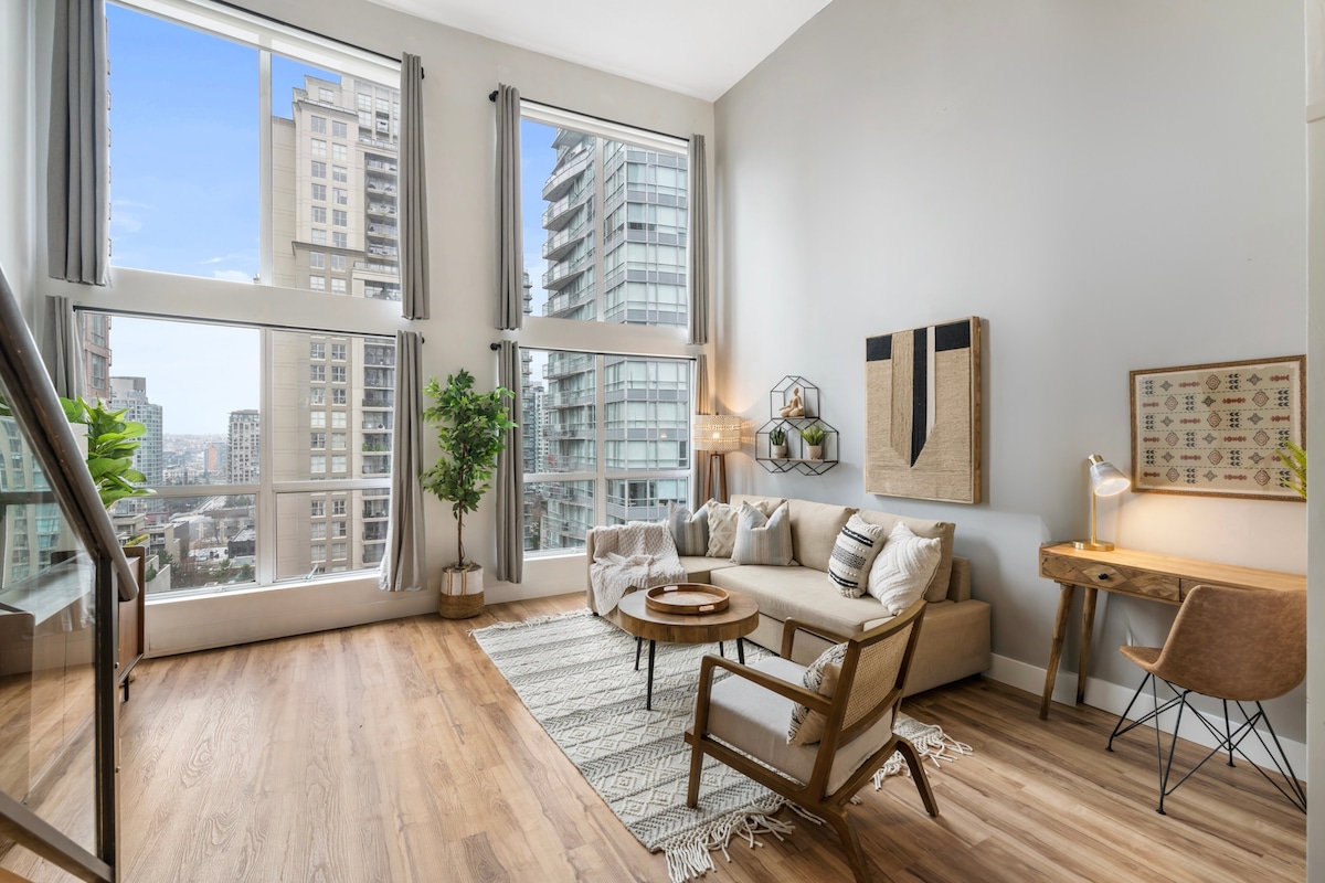 A spacious living area is presented with floor-to-ceiling windows offering views of the cityscape. Natural light fills the room, highlighting a comfortable couch and a round coffee table. A small workspace with a lamp is included, along with a decorative plant and modern accent chair.