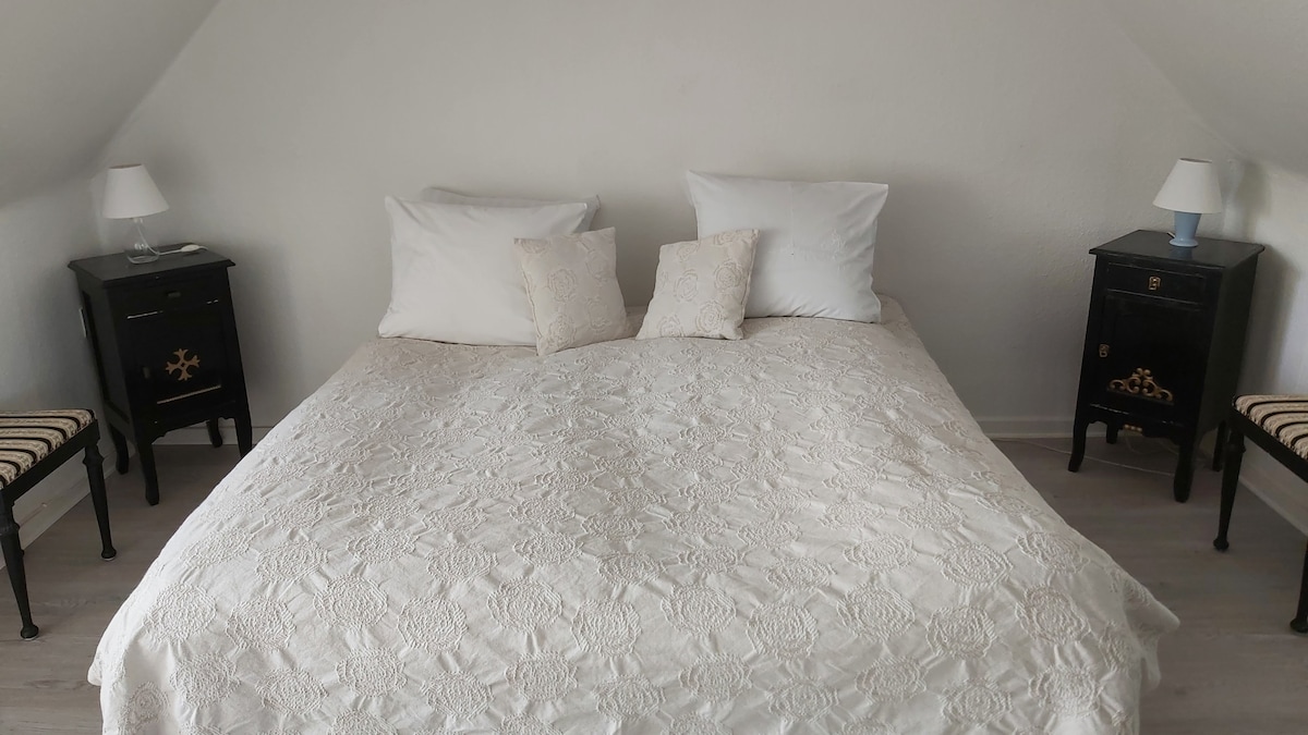 A cozy bedroom features a spacious double bed adorned with a textured white bedspread and multiple pillows. Two bedside tables with lamps are positioned beside the bed, complemented by two simple chairs at the foot.