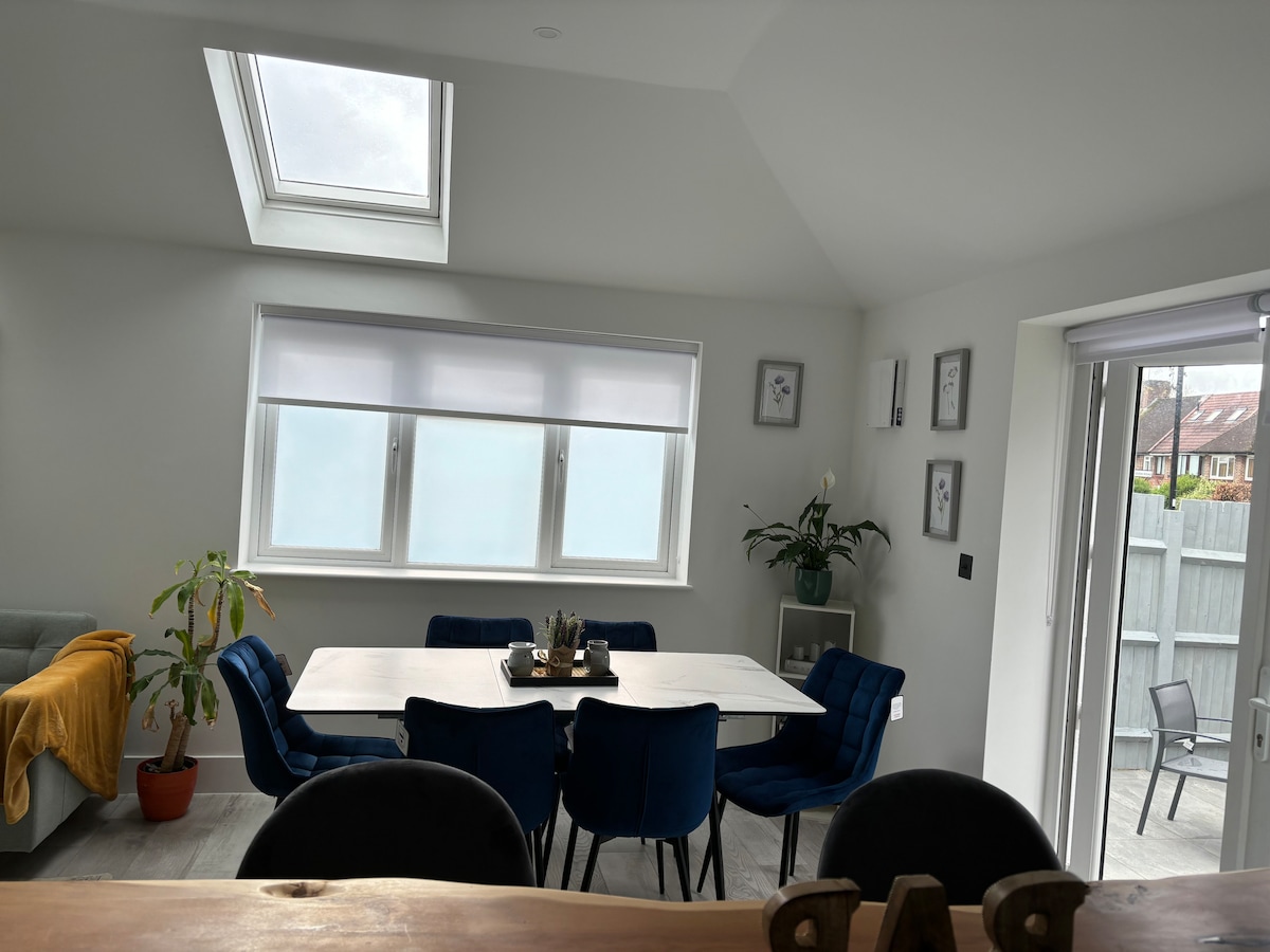 A modern dining area is highlighted by a large, rectangular table surrounded by six blue chairs. Natural light fills the space through a skylight and two windows. A potted plant adds a touch of greenery, while patio doors lead to the garden area.