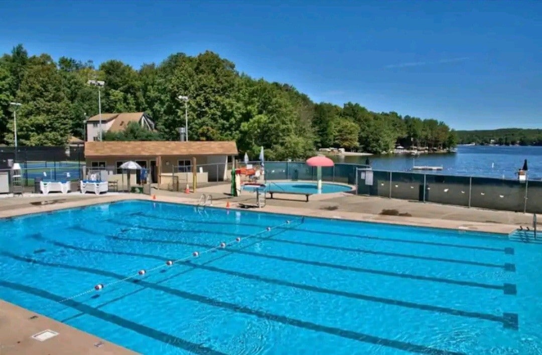 An outdoor swimming pool is seen with clear blue water, lined by lounge chairs and umbrellas. In the background, a serene lake reflects the surrounding greenery, while a spacious deck offers additional seating. The area is well-lit, providing a welcoming atmosphere for relaxation.