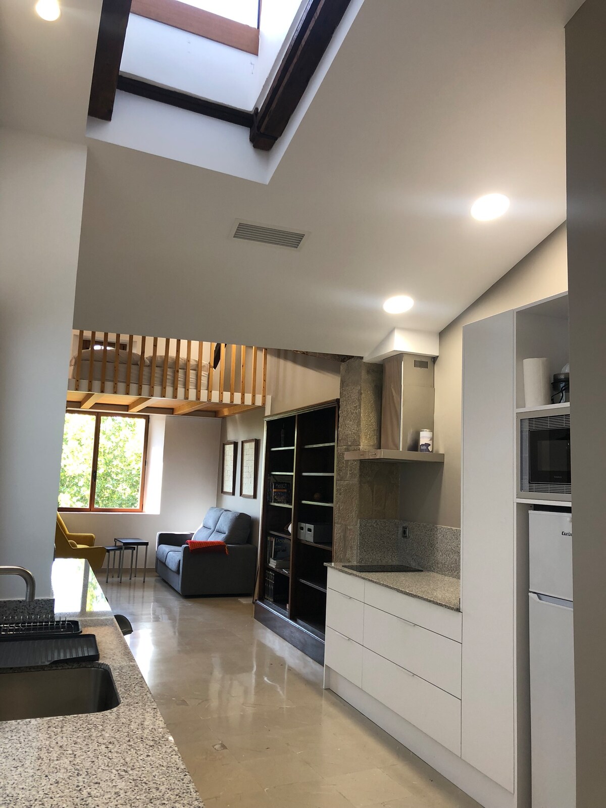 An open-plan kitchen area is visible, showcasing modern cabinetry and a granite countertop. Natural light filters through a skylight and large windows, illuminating the space. A small seating area with a grey sofa and a bright armchair can be seen in the background, enhancing the inviting atmosphere.
