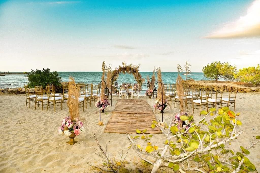 A beautifully arranged beach wedding setup features golden chairs aligned on soft sand. An elegantly crafted arch adorned with dried florals frames the ocean view, while flower arrangements add color to the scene. The serene water and clear sky create a tranquil atmosphere.