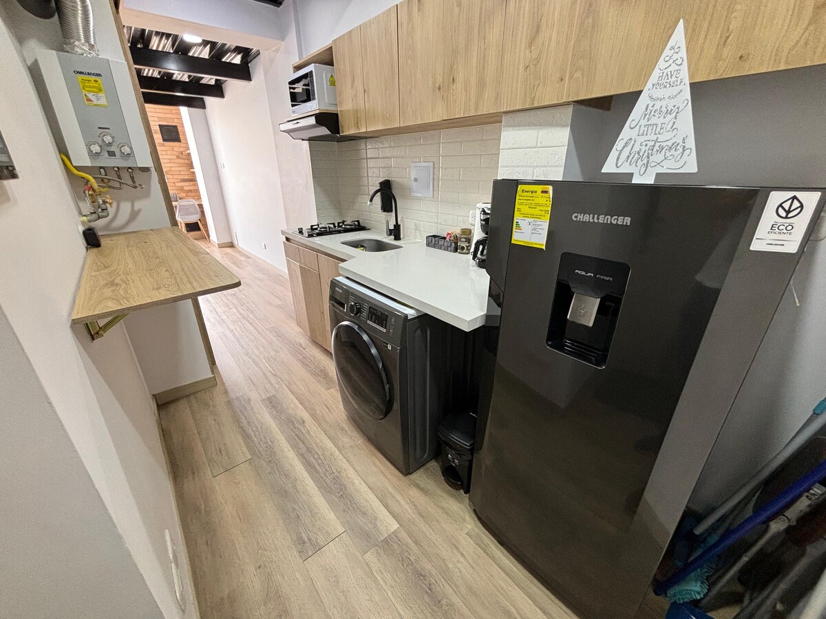 A compact kitchen area is presented with modern appliances, including a refrigerator, washing machine, and stovetop. Light wooden cabinetry complements the clean, white countertops. Wooden flooring extends throughout the space, contributing to a functional and efficient layout.