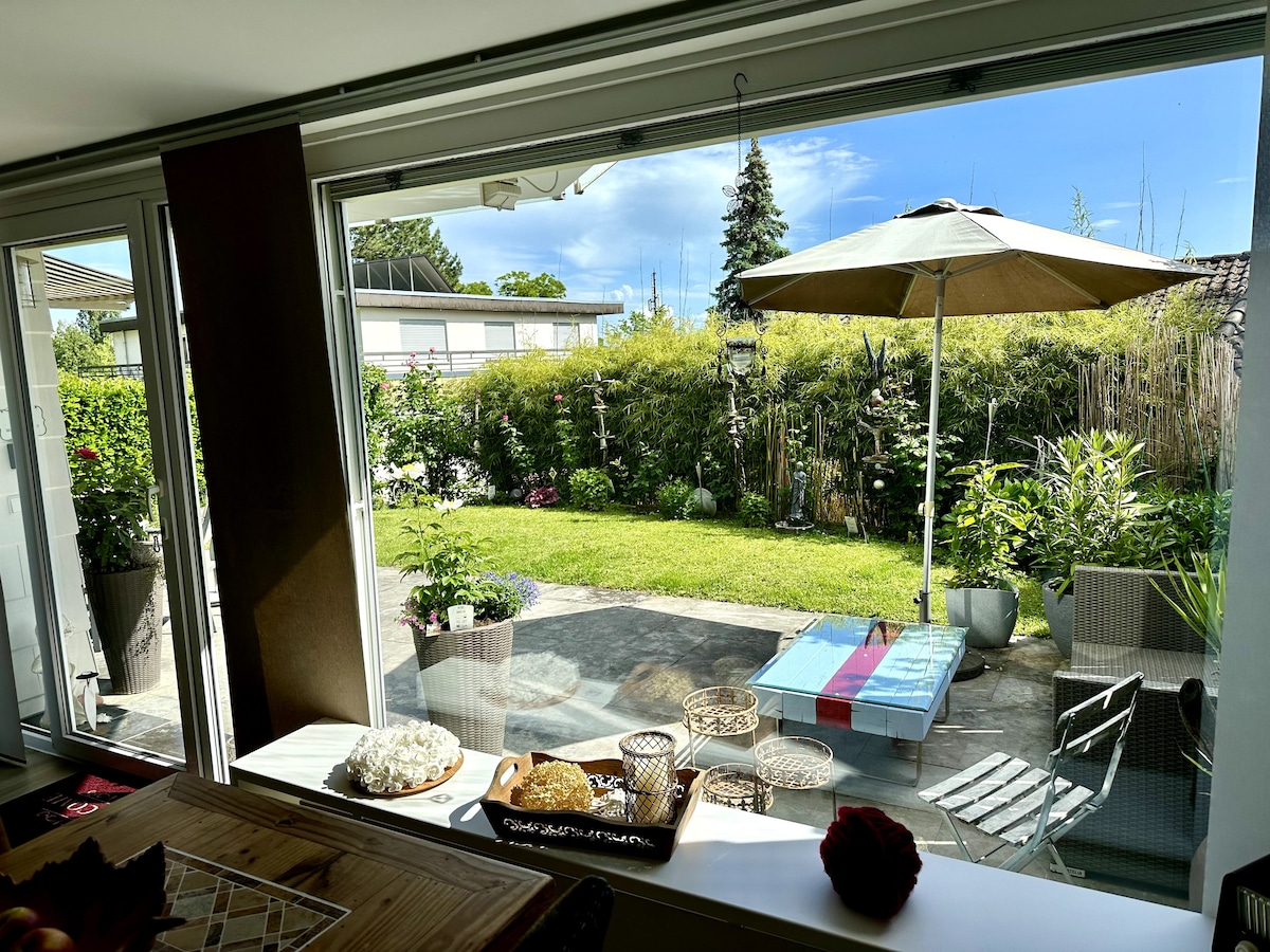 A spacious window frames a serene garden view, featuring lush greenery and a patio with an umbrella. A low table is set with a selection of baked goods, while comfortable seating arrangements provide a welcoming space for relaxation.