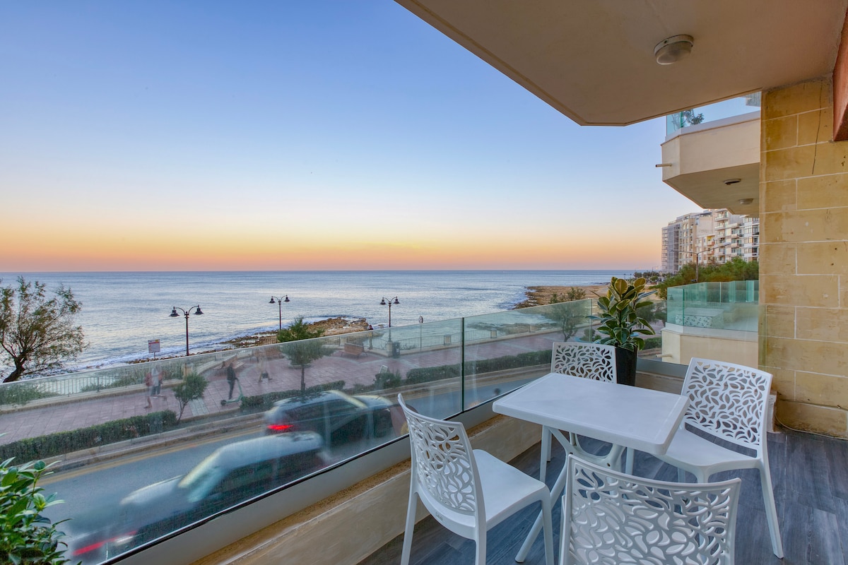 A spacious balcony overlooks the sea, featuring modern outdoor furniture with white, intricately designed chairs and a matching table. The horizon glows with warm colors as the sun sets, creating a serene coastal view.