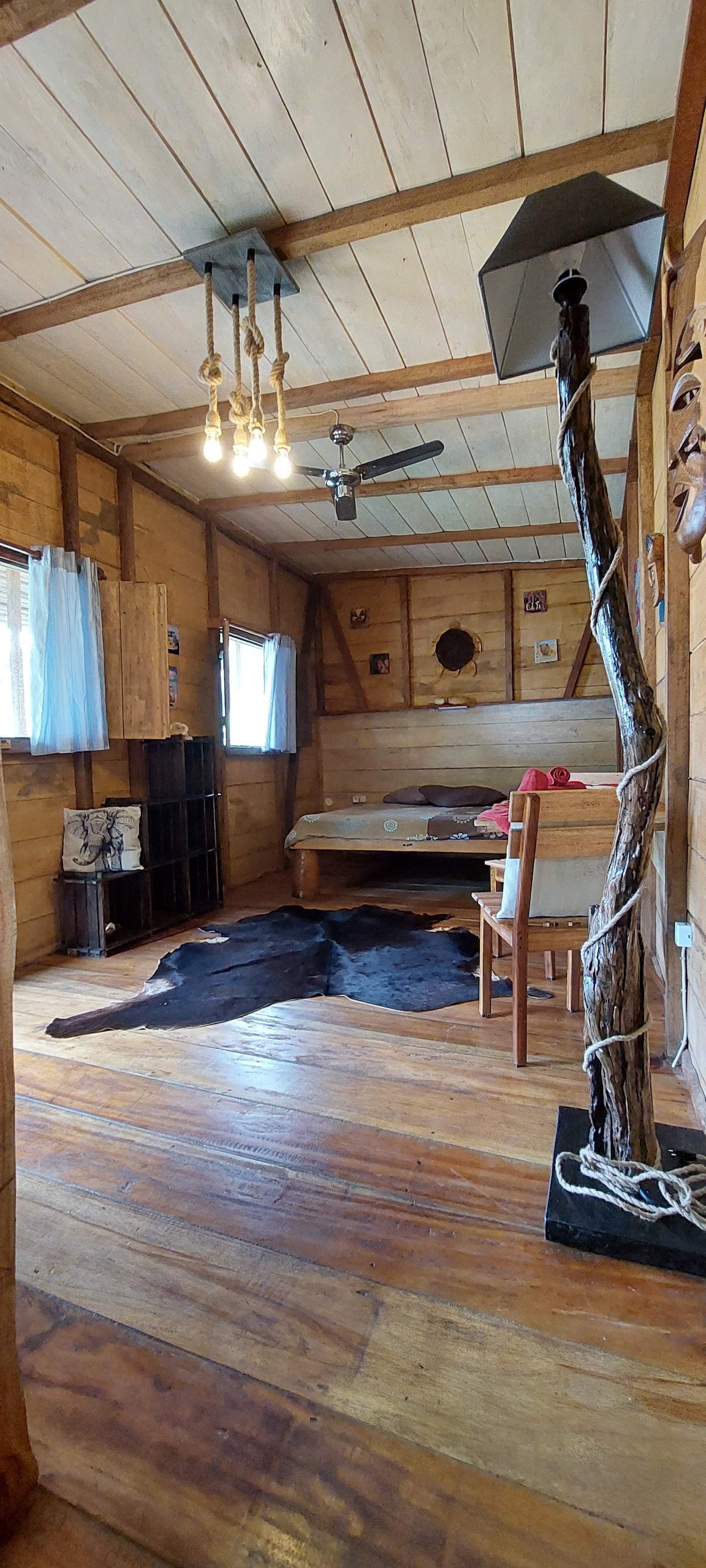 The interior of the bungalow features wooden walls and ceiling, complemented by warm wooden flooring. A spacious bed is situated at the back, while a small dining area is to the side. A unique lamp and rustic decor add character to the room, which is well-lit by natural light from the windows.