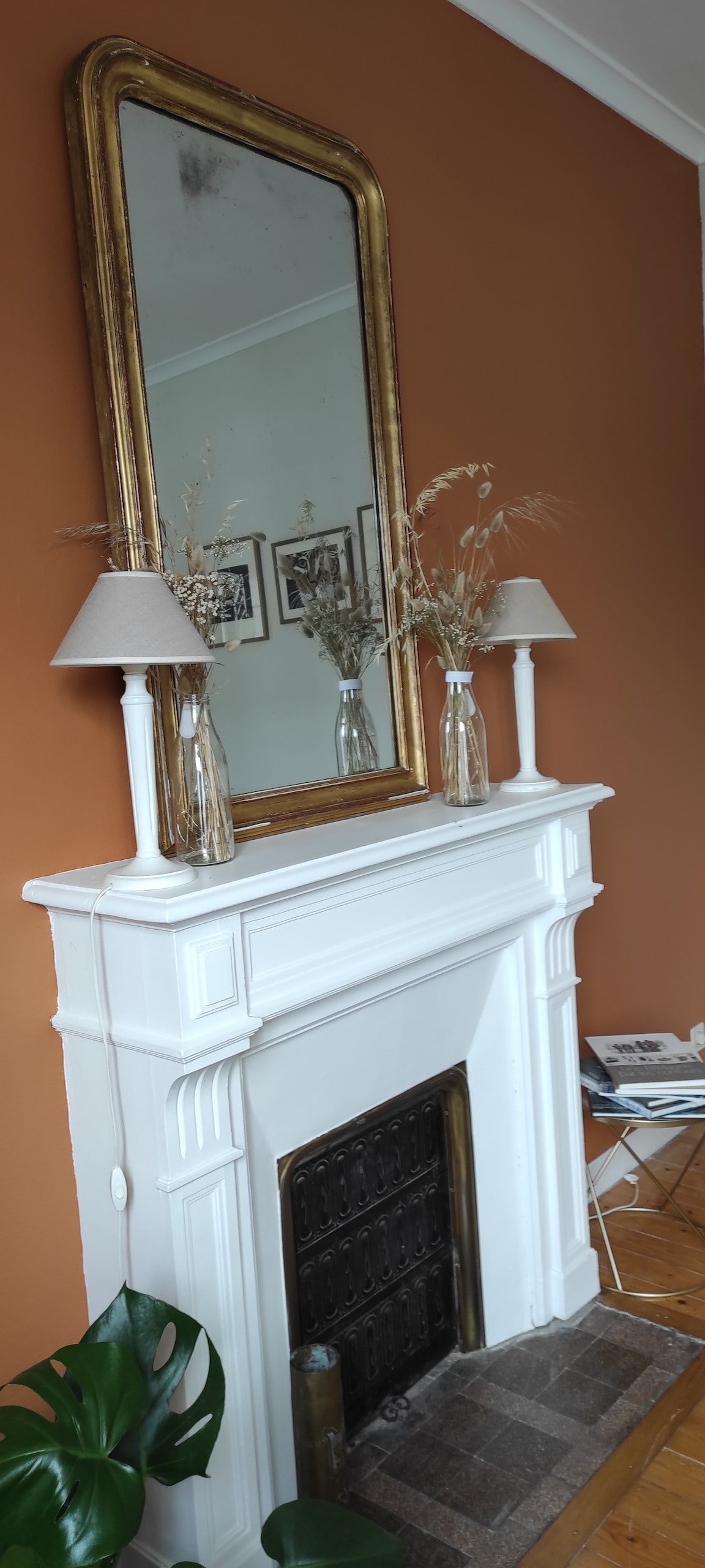 A white mantelpiece features an ornate design and is topped with a large mirror framed in gold. Two neutral-toned lamps are positioned on either side, accompanied by slender vases containing dried flowers. The backdrop is complemented by a warm, terracotta wall.