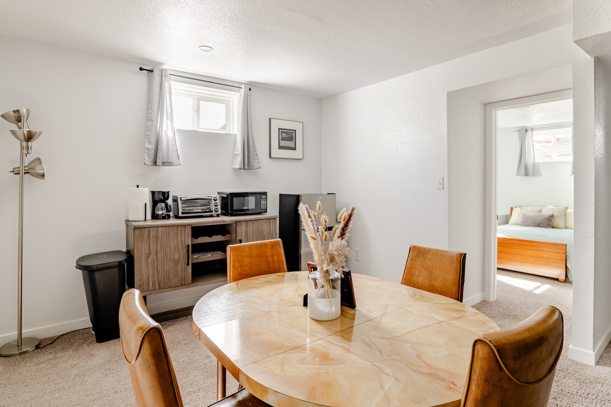 A well-lit kitchenette area features a round dining table surrounded by four leather chairs. A sleek wooden cabinet houses a microwave, refrigerator, and a coffee maker. Light filters in through a window dressed with light grey curtains, providing a cozy ambiance.