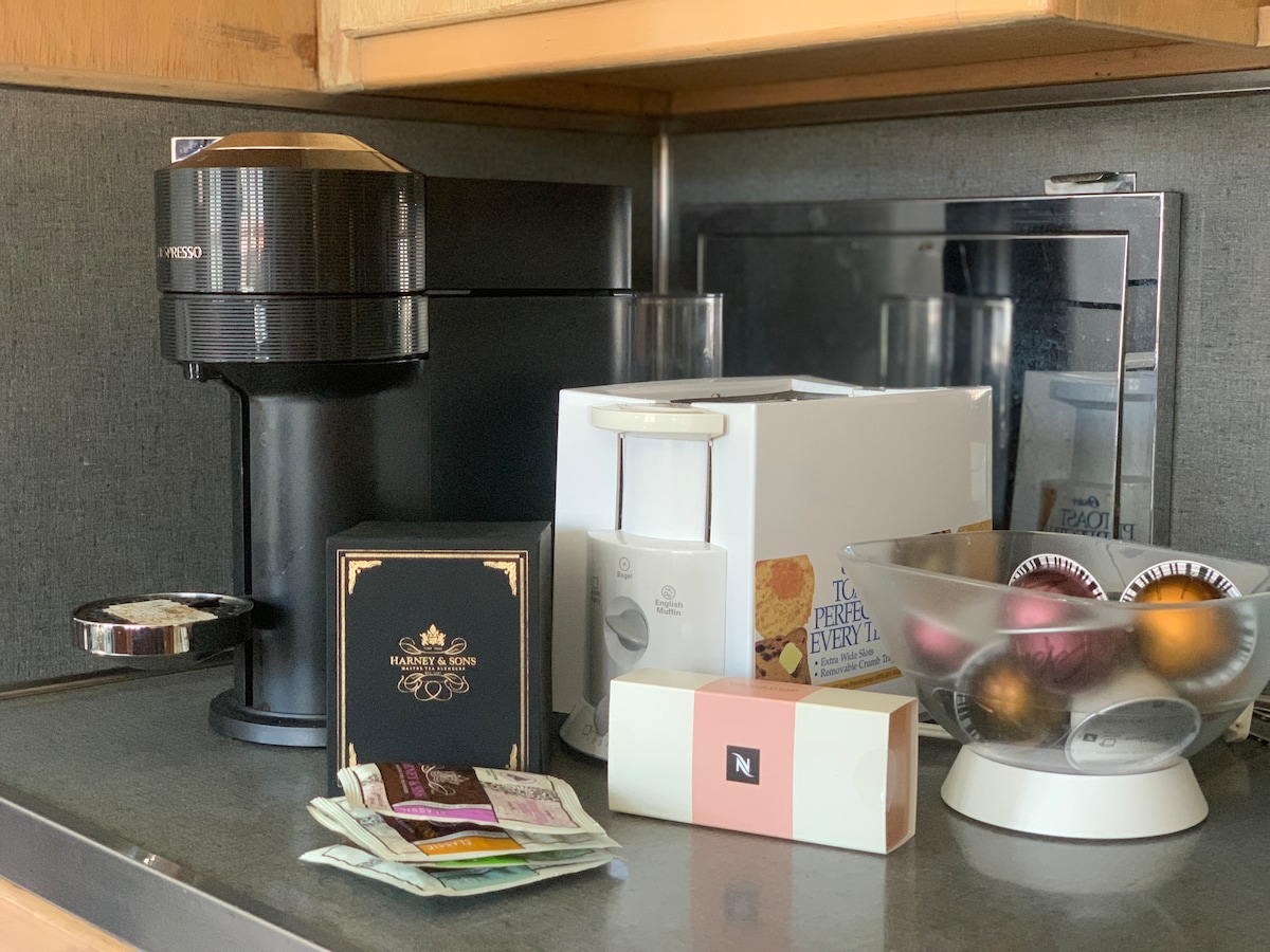 A sleek coffee machine sits on a countertop beside a compact toaster and a decorative bowl filled with colorful coffee pods. Various packets of tea and coffee options are arranged nearby, providing a selection for guests. A microwave is positioned in the background.