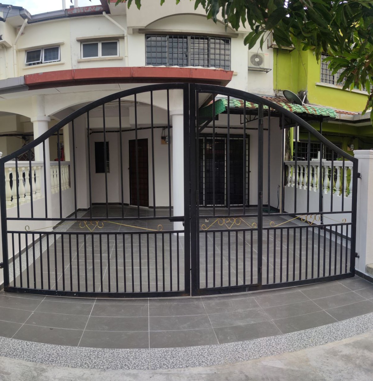 A black, wrought-iron gate opens into a tiled entrance area, showcasing a clean and spacious outdoor patio. The area is bordered by white walls and features decorative elements near the entrance. Nearby greenery enhances the inviting atmosphere.