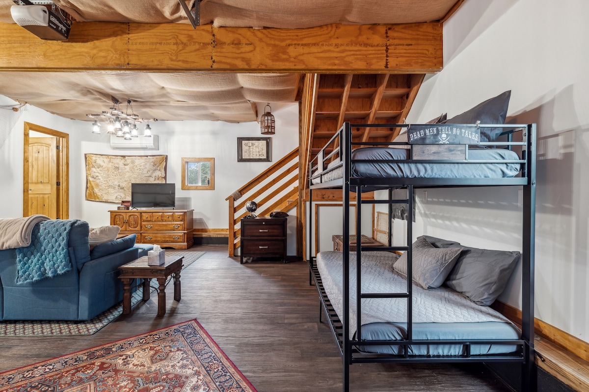 A spacious loft area features a cozy couch paired with a patterned area rug. A set of twin bunk beds is positioned against the wall, with a light-colored mattress on each level. The room’s wooden beams and soft lighting create a warm environment.
