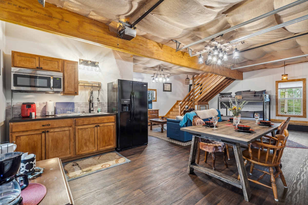 A bright and airy kitchen area features wooden cabinetry and modern appliances, including a microwave and refrigerator. A dining table with wooden chairs is positioned near the kitchen. Natural light enters through windows, illuminating the space with a relaxed ambiance.