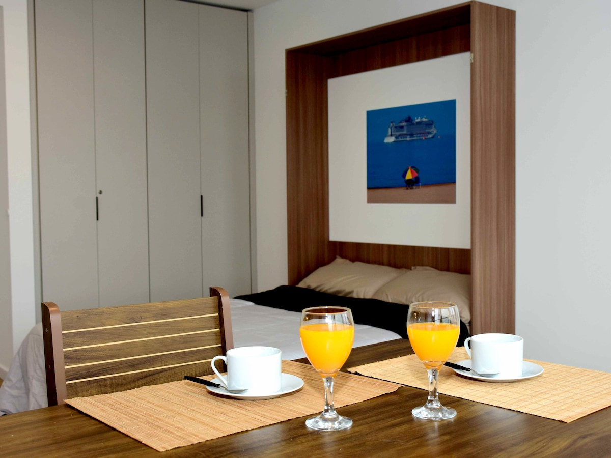 A dining table is set with two cups and glassware filled with orange juice. A spatial wall bed is positioned in the background, complemented by neutral-toned bedding. Pared-down cabinets add functionality to the room, while a tranquil photo of a beach scene enhances the ambiance.