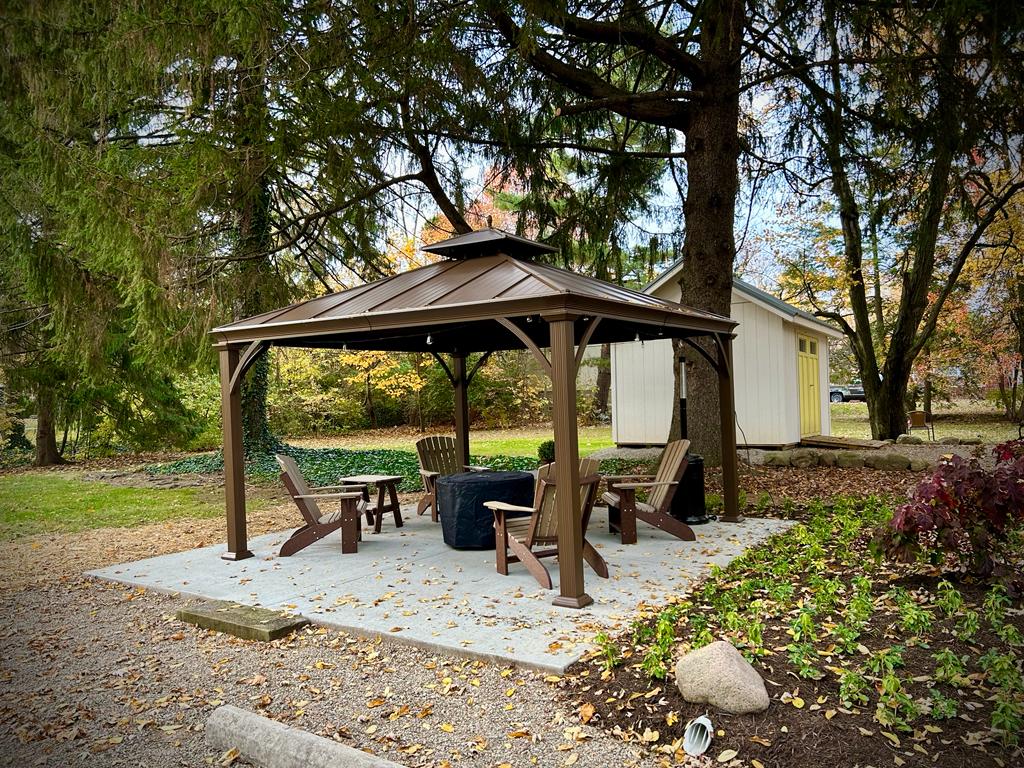 A covered gazebo is situated on a stone patio, surrounded by trees. Several wooden Adirondack chairs are arranged around a small table. Autumn leaves scatter across the ground, and a storage shed is visible in the background, enhancing the outdoor gathering space.