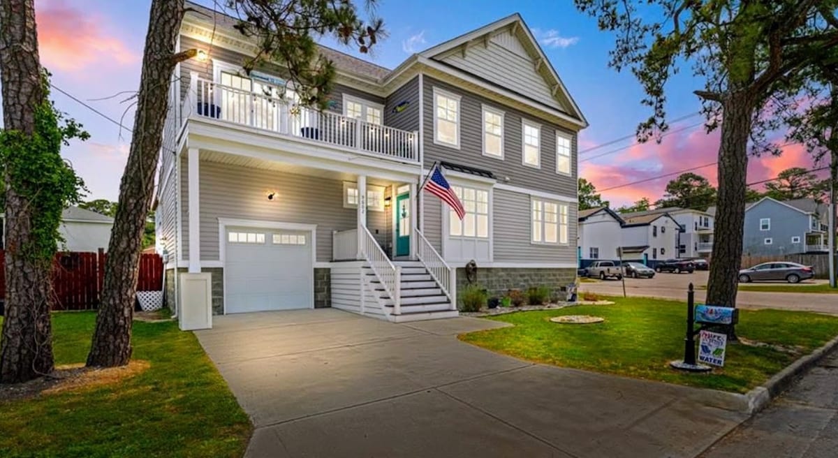 The exterior of the home features a welcoming porch with a railing, flanked by trees and a neatly trimmed lawn. An American flag is displayed prominently. The driveway leads to a garage, and the overall structure showcases a modern design with light-colored siding.