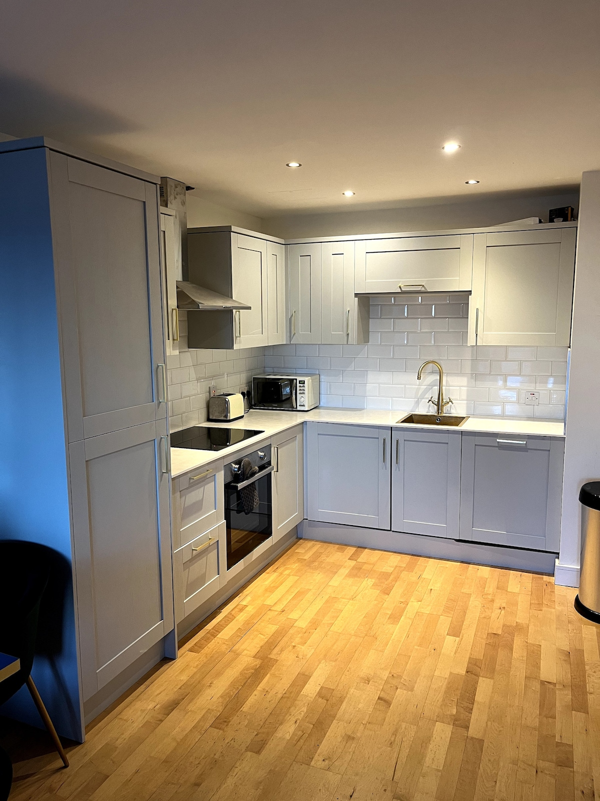 A modern kitchen features grey cabinetry with a white tiled backsplash. Appliances include a microwave, a washing machine, and a dishwasher. A stainless steel sink is centrally located, illuminated by recessed lighting above. The floor is composed of light wooden planks, contributing to a clean, contemporary look.