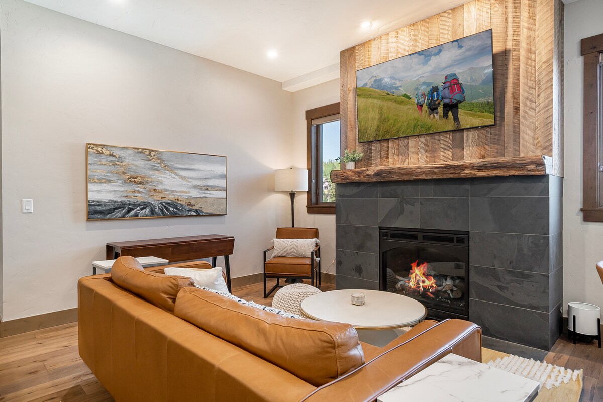 A modern living area features a cozy leather sofa facing a sleek gas fireplace. A large TV mounted above the fireplace displays a scenic mountain view. Natural light filters through the nearby windows, enhancing the warm tones of the wood accents.