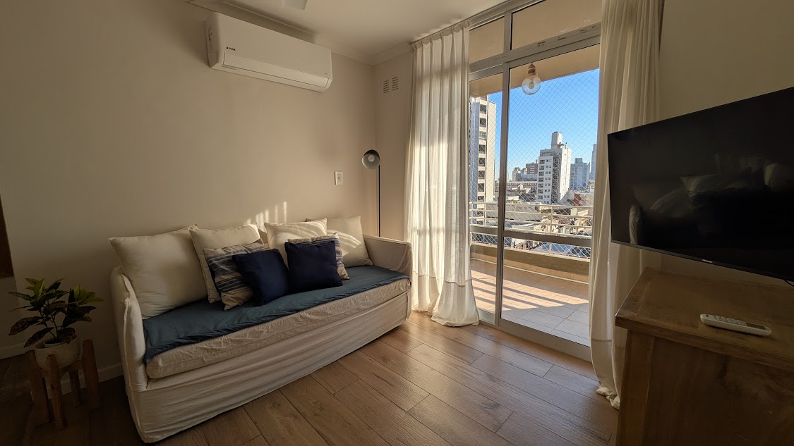 A bright living space features a light-colored sofa adorned with blue and neutral cushions. Natural light floods in through spacious glass doors that open to a balcony, while a flat-screen TV is positioned across from the seating area. Light-colored wooden floors enhance the clean aesthetic.