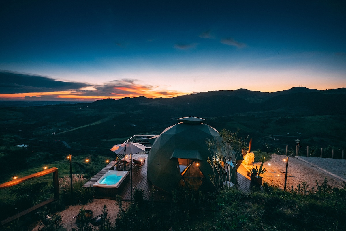 A geodesic dome is set upon a deck overlooking rolling hills at twilight. A small hot tub is positioned alongside the dome, illuminated by soft lighting. The expansive sky displays warm hues of sunset, enhancing the tranquil ambiance of this unique getaway.