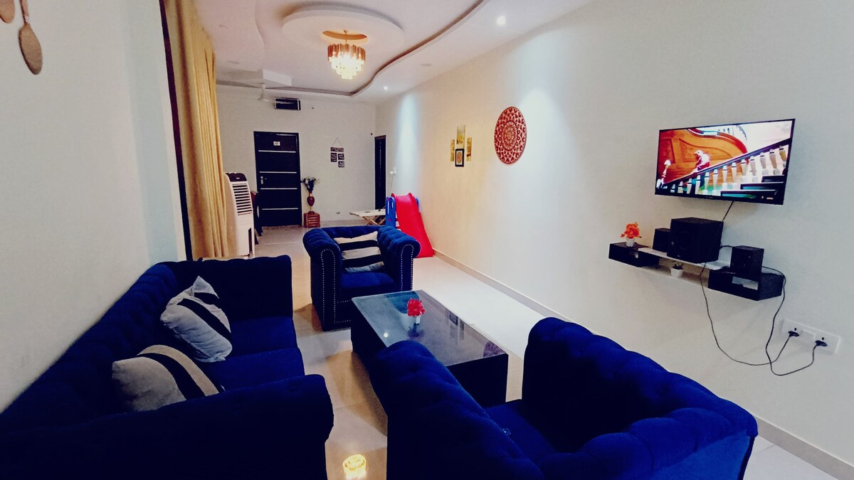 The living area features a collection of deep blue sofas arranged around a glass coffee table. A television is mounted on the wall, and decorative elements add character to the space, which is illuminated by soft ceiling lights.