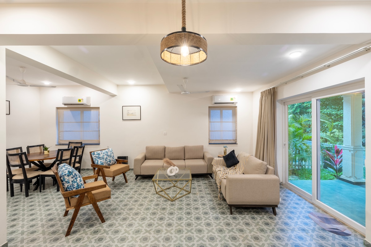 The living area features a spacious layout, showcasing a light-coloured sofa set and a glass coffee table. Two accent chairs with vibrant cushions are positioned nearby. A dining table is visible in the corner, accompanied by stylish black chairs. Large sliding doors lead to a garden view.