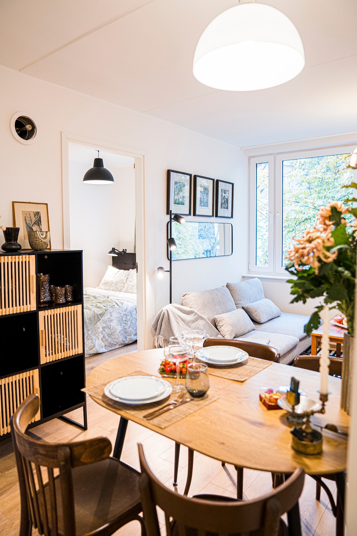 A small dining area features a wooden table set for two, with plates and glassware in place. A cozy sitting area with a light sofa is visible in the background, along with natural light streaming through large windows. Decorative elements are present on the table and walls.