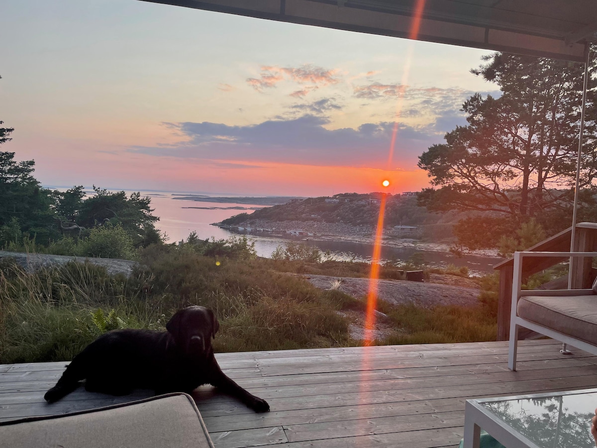 A peaceful outdoor area features a wooden deck with a spacious seating arrangement. In the foreground, a black dog is resting on the deck, while the background showcases a vibrant sunset over a tranquil coastline, highlighting the natural beauty of the surroundings.