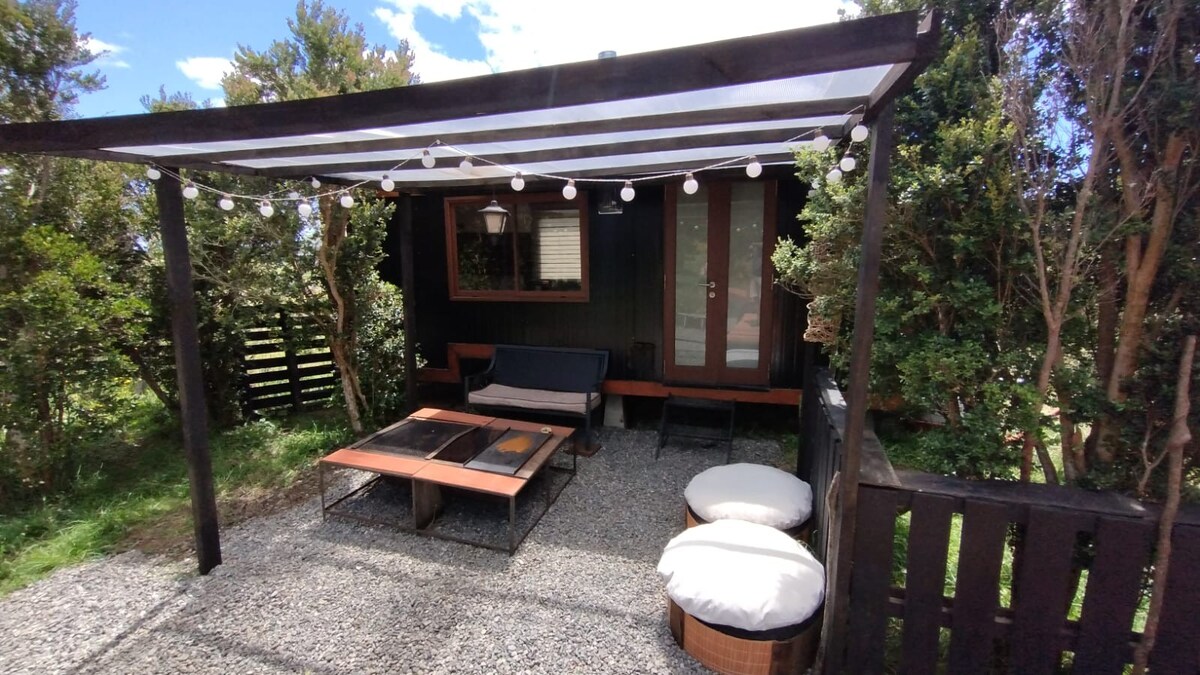 An outdoor seating area features a shaded pergola with string lights overhead. A low table sits in the center, surrounded by a cozy sofa and oversized cushions. Lush greenery encloses the space, creating a sense of privacy and connection to nature.