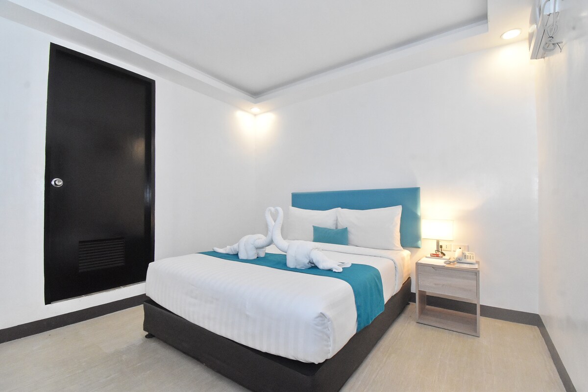 A compact room features a plush Queen bed adorned with soft linens and decorative pillows. Elegant towel sculptures are displayed on the bed. A bedside table holds a lamp, and a window allows natural light to brighten the white and teal color scheme, enhancing the tranquil ambiance.