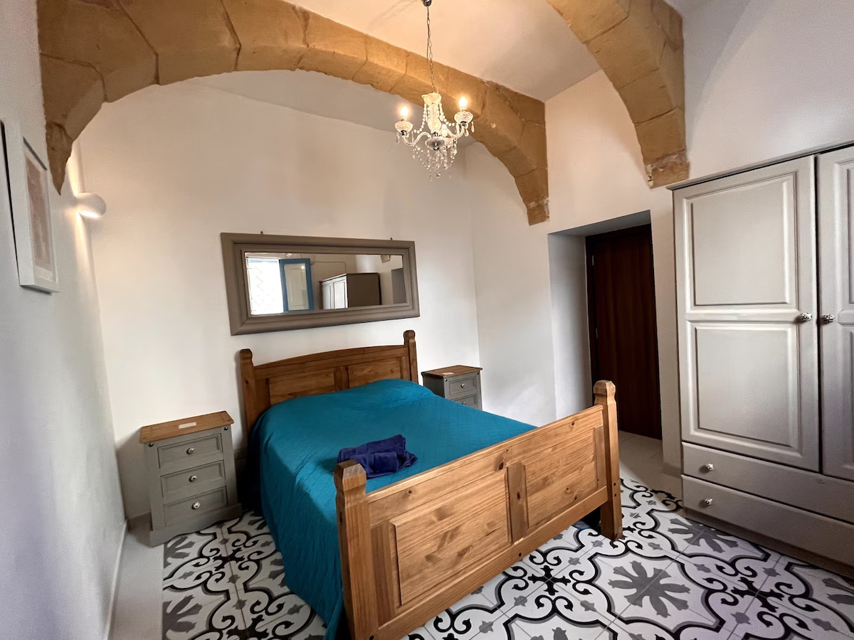 The spacious sleeping area features a wooden bed with a blue coverlet, flanked by two bedside tables. A large mirror hangs above the bed, reflecting the room's high arched ceiling, which is accented by stone detailing. A wardrobe is positioned against one wall.