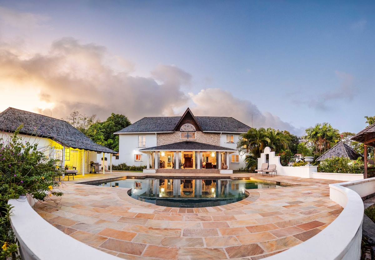 The villa's exterior is showcased, featuring a spacious pool surrounded by natural stone tiles. Lush greenery and tropical plants frame the yard, while the picturesque architecture blends harmoniously with the landscape under a soft twilight sky.