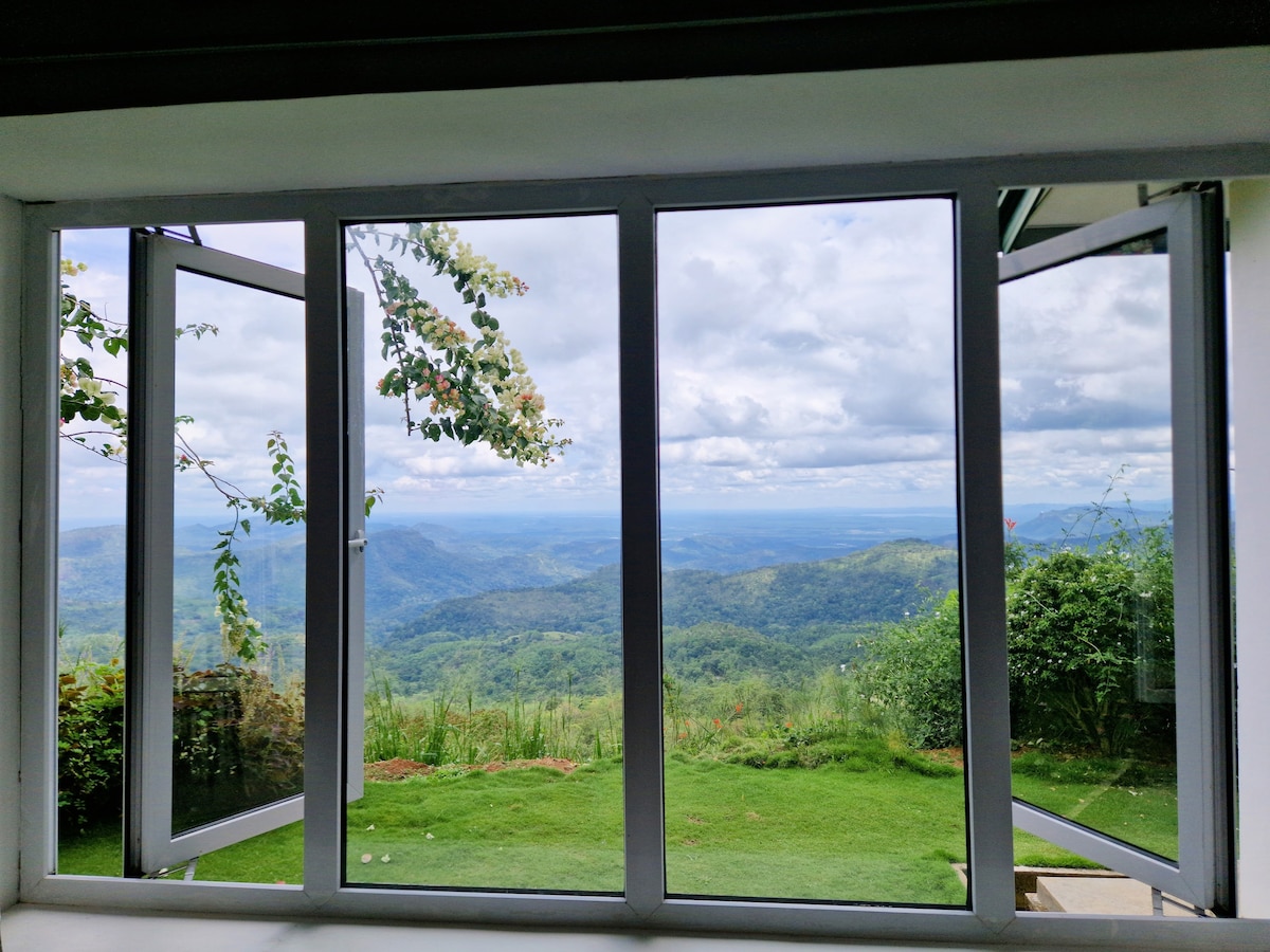 A spacious window with glass panes that open wide provides a view over lush green hills and valleys. The landscape stretches far into the distance, offering a tranquil setting with cloud-covered skies overhead. Garden foliage frames the view outside.