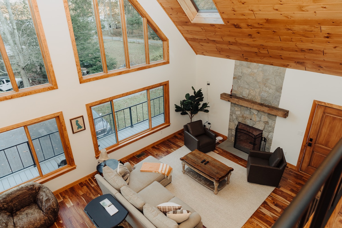 A spacious living area is highlighted by a high wood-paneled ceiling and large windows allowing natural light. A stone fireplace is centrally located, complemented by comfortable seating and a wooden coffee table on a light-colored rug.