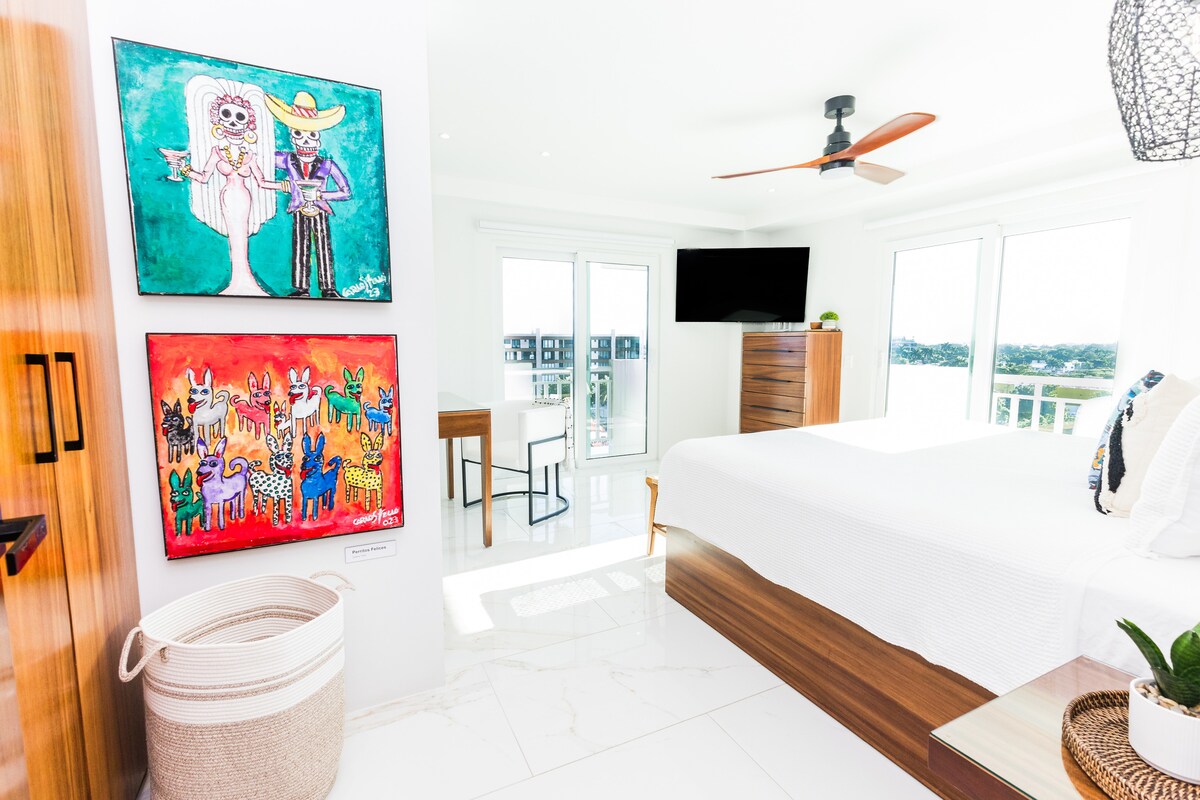 A bright and airy bedroom features large windows providing natural light and beautiful views. Local art adorns the walls, with colorful pieces displayed alongside a minimalist writing desk. Modern furnishings, including a spacious bed and a sleek chest of drawers, complement the airy ambiance.