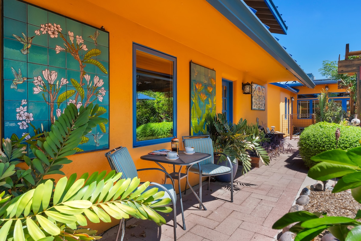 A vibrant, sunlit patio area showcases colorful wall art on bright orange walls. A small table with two chairs sits beneath lush greenery, providing a perfect spot for outdoor dining. The space is adorned with various plants, enhancing the inviting atmosphere.