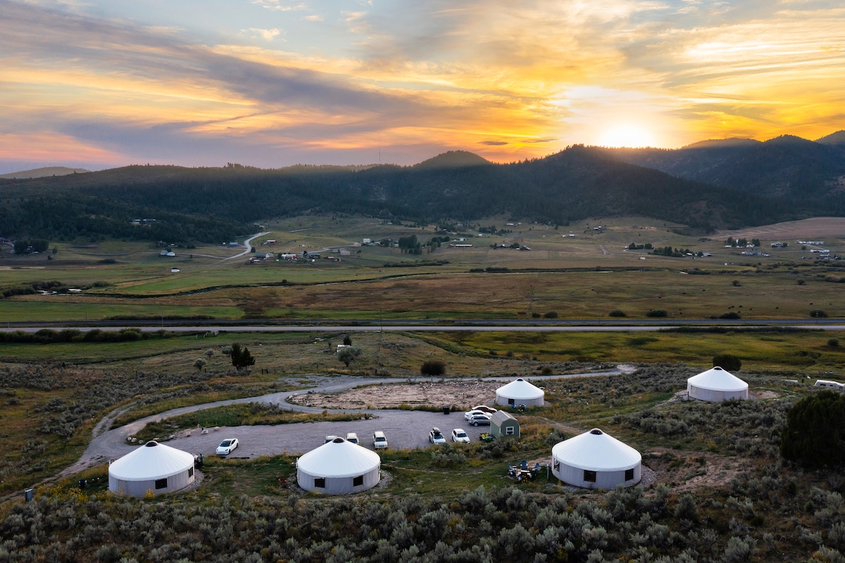 A panoramic view captures the serene hillside setting at sunset, showcasing five circular yurts arranged in a circular layout. Surrounding fields and gentle mountains create a calming backdrop, while the colorful sky reflects a blend of warm hues, indicating the end of the day.